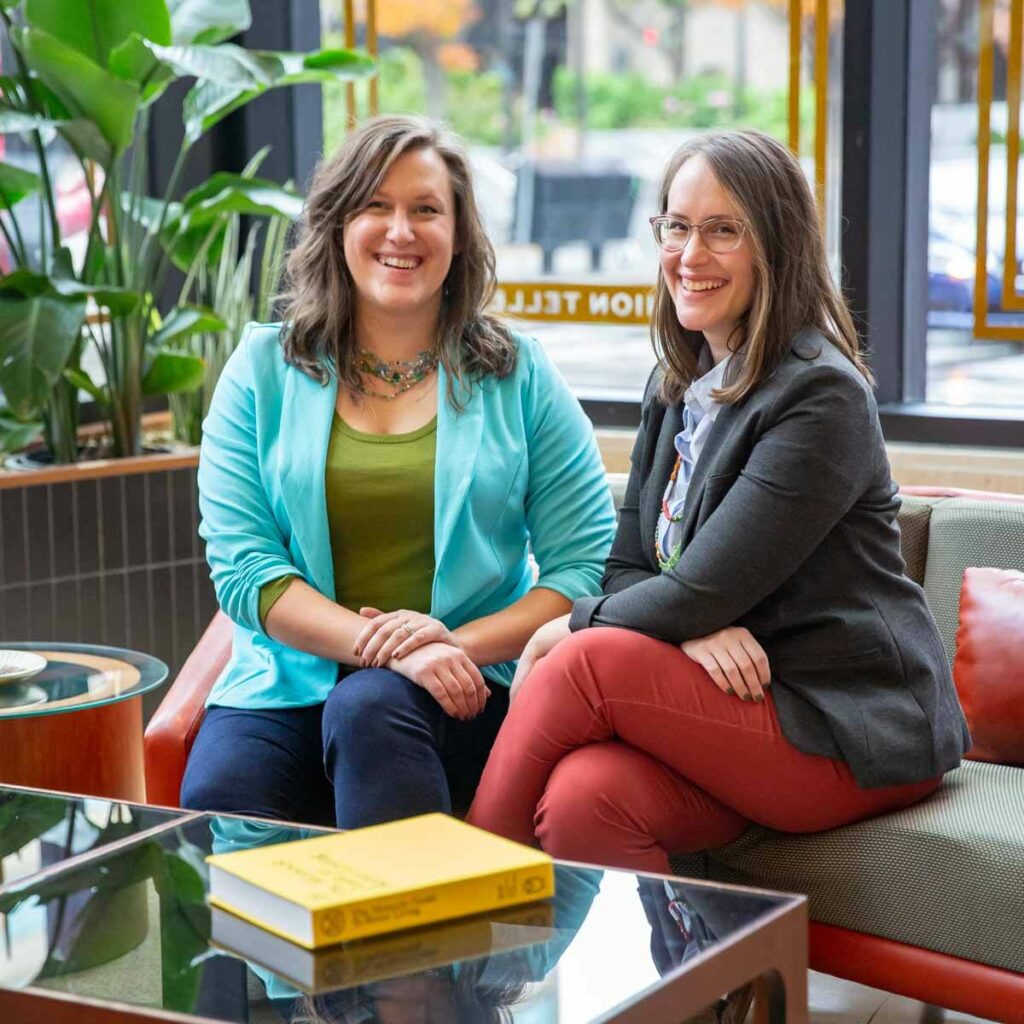 Jessica Yaeger and Christina Blust, founders of Blustery Day Design, seated on a sofa in front of a window.