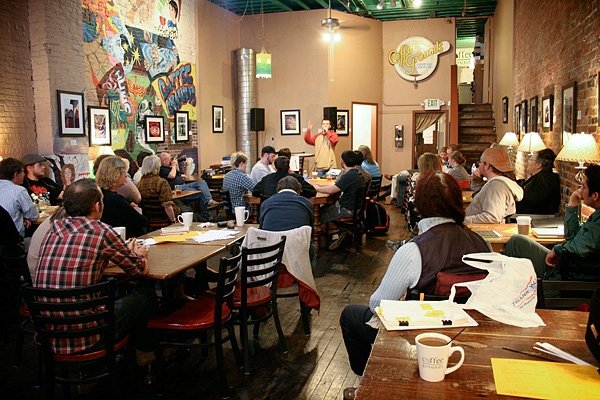 Various people sitting at tables at a coffee shop, listening to someone reading a poem at the podium.