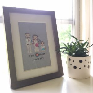 A framed cross stitch portrait of a family, stitched on a mid-gray aida cloth.