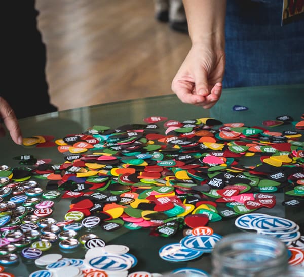 A hand picking up a branded guitar pic from a colorful spread of pics on the table.