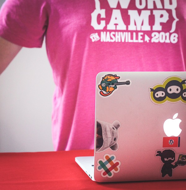 A man in a pink WordCamp t-shirt next to a laptop with the Wapuu sticker on its cover