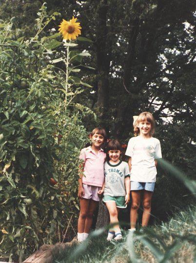 Three young girls smiling next to a lovely yellow sunflower towering over them in their childhood backyard.